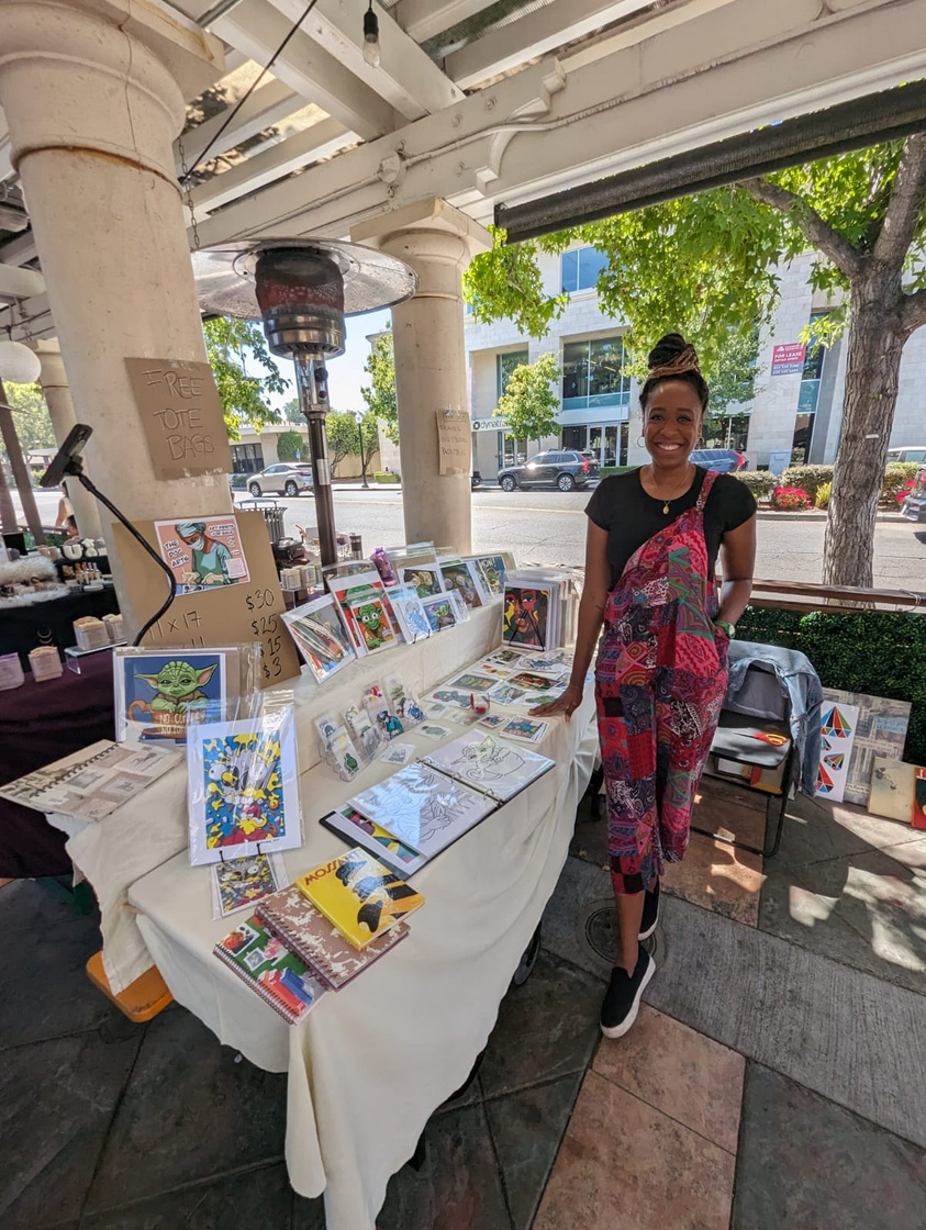 Photo of the artist next to her display of all of her arts at her first vendor event. She is wearing colorful overalls and a black shirt. 